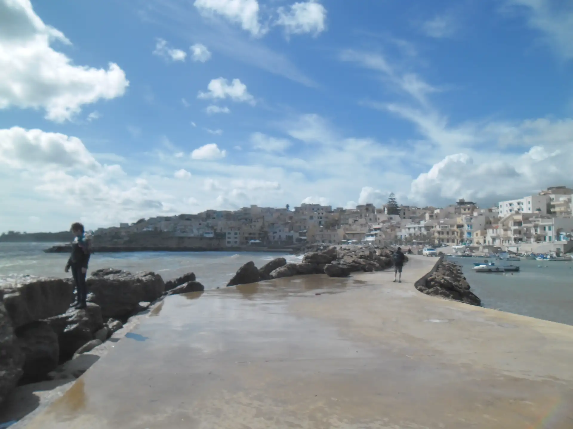 向海心延伸的西侧防波堤（Molo di Sopraflutto）。沿着长长的码头栈道漫步，视野极为开阔。你可以看到渔民们在此驻足垂钓或修补渔网。由此向北回望，可以将整座渔村的白色建筑轮廓线以及远处山丘上雄伟的古希腊神庙遗迹同时定格在镜头中。